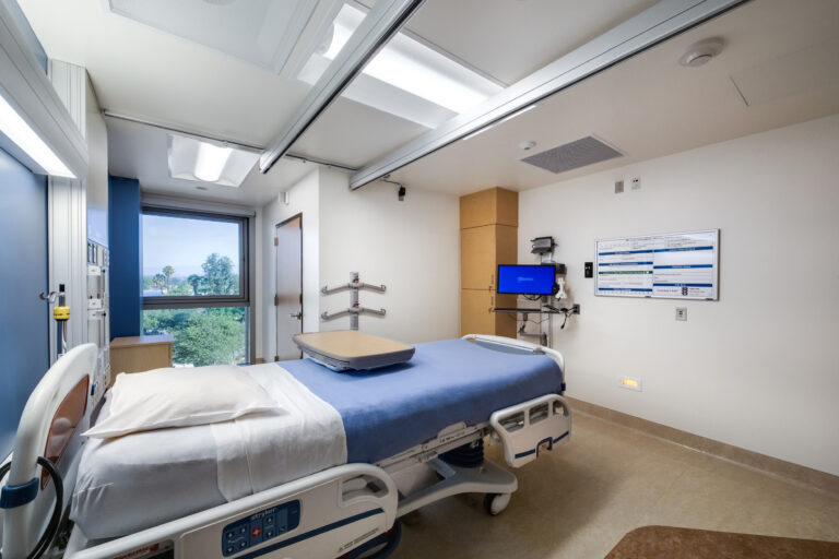 Modern hospital room with an empty patient bed, wall-mounted monitor, and large window overlooking trees