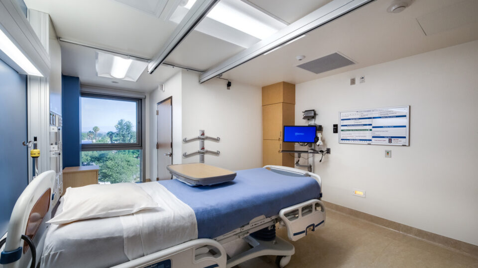 Modern hospital room with an empty patient bed, wall-mounted monitor, and large window overlooking trees