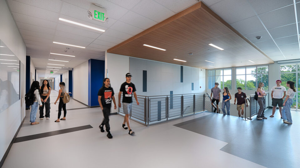 Students walking and talking in a modern school hallway with large windows and an exit sign overhead