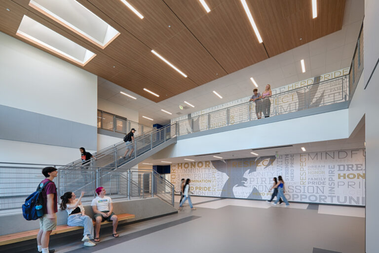 Modern school atrium with students walking, sitting, and chatting beneath a wood-paneled ceiling and staircase