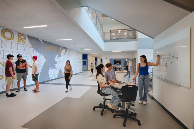Students socialize and study in a bright modern school hallway, with a teacher writing on a whiteboard