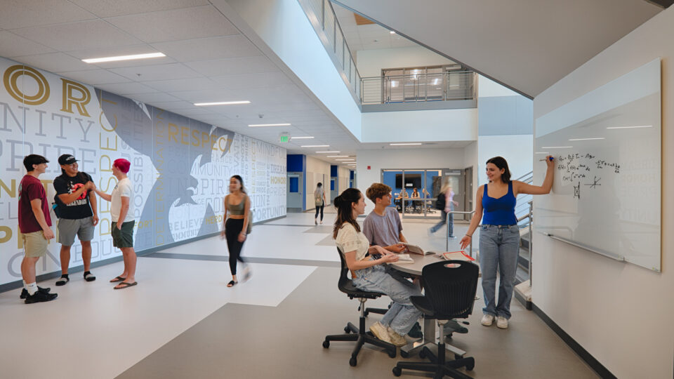 Students socialize and study in a bright modern school hallway, with a teacher writing on a whiteboard