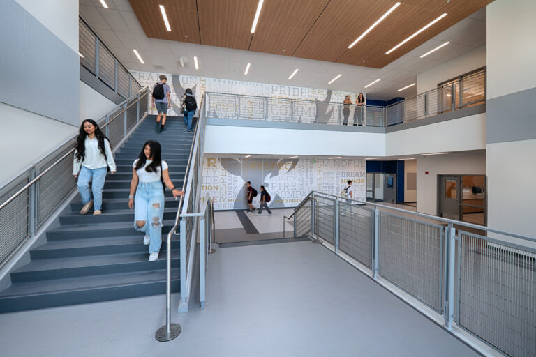 Students walking through a bright modern school atrium with stairs, railings, and wall words about pride and respect