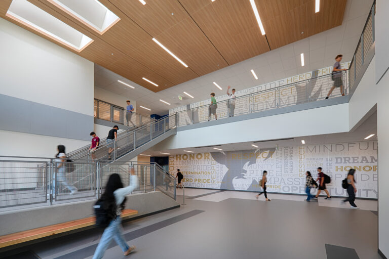 Students walking through a modern school atrium with staircases, railings, and a large motivational mural