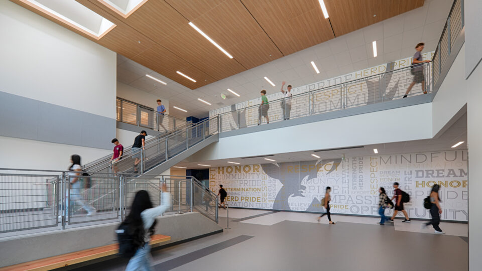 Students walking through a modern school atrium with staircases, railings, and a large motivational mural