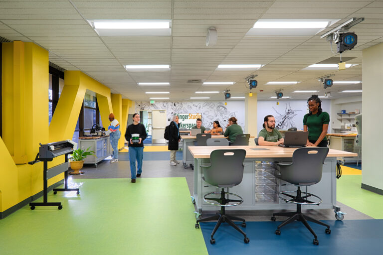 People working in a bright makerspace with 3D printers, worktables, and yellow wall accents