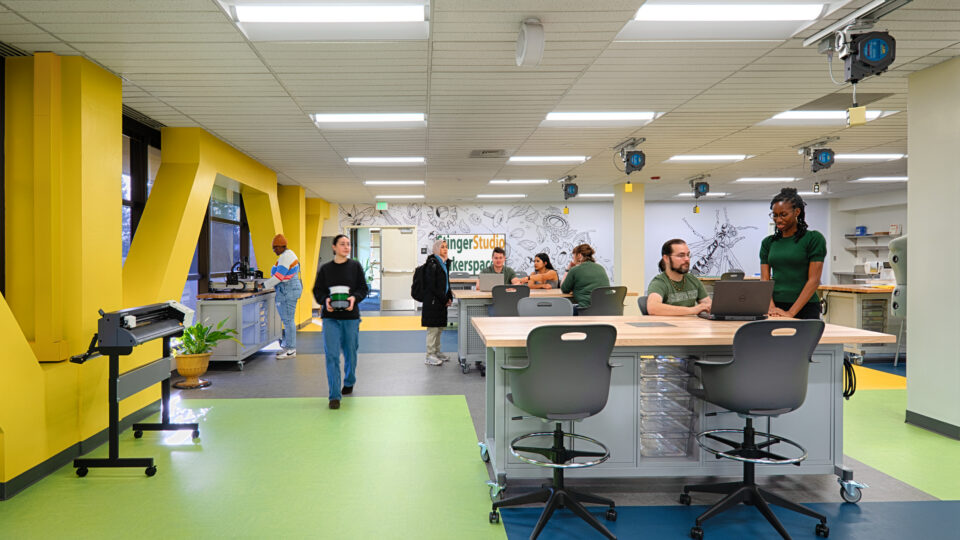 People working in a bright makerspace with 3D printers, worktables, and yellow wall accents