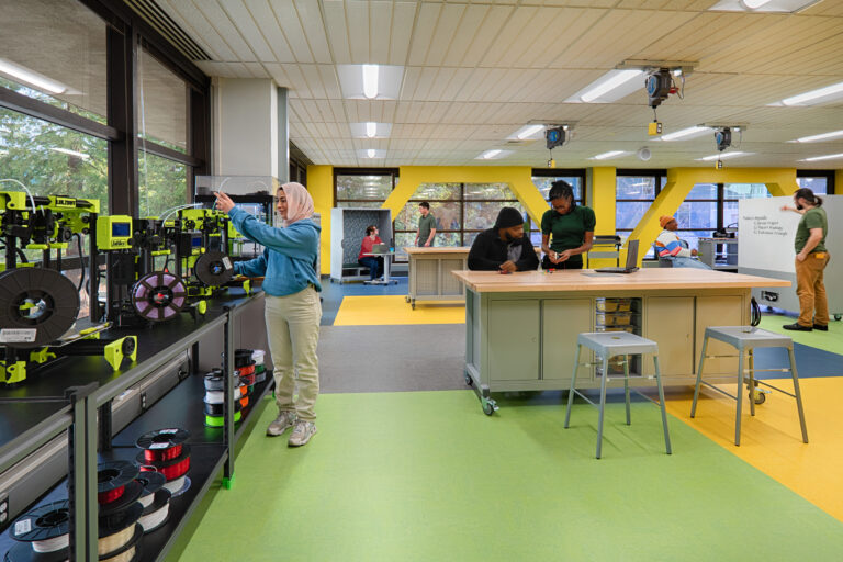 People working in a bright makerspace with 3D printers, a central workbench, and a whiteboard agenda