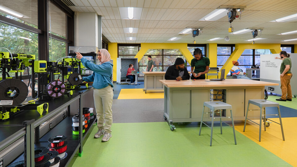 People working in a bright makerspace with 3D printers, a central workbench, and a whiteboard agenda