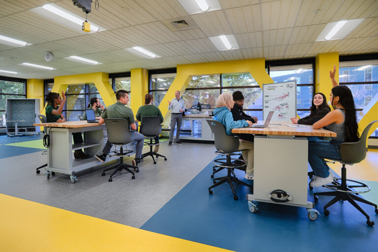 Students in a modern classroom raise hands during a collaborative lesson with a teacher at the front