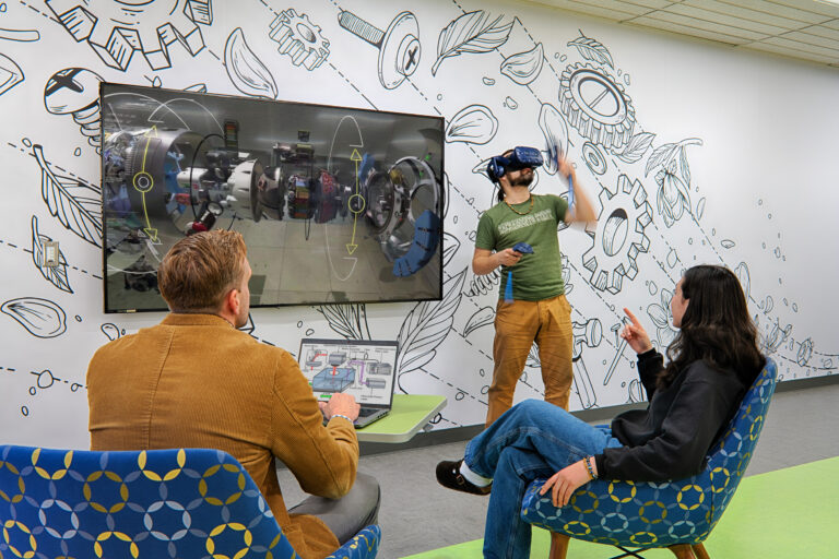 People in a modern meeting room watch a man demonstrate VR beside a large screen showing a 3D technical model