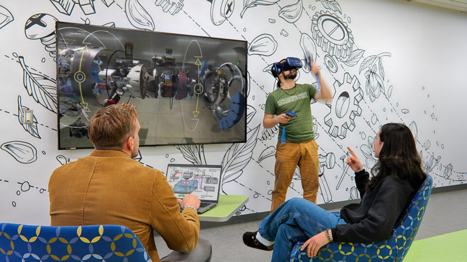 People in a modern meeting room watch a man demonstrate VR beside a large screen showing a 3D technical model