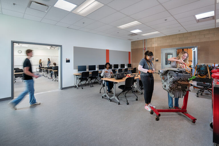 Students in a classroom lab work on a large engine while others study at computers in the background