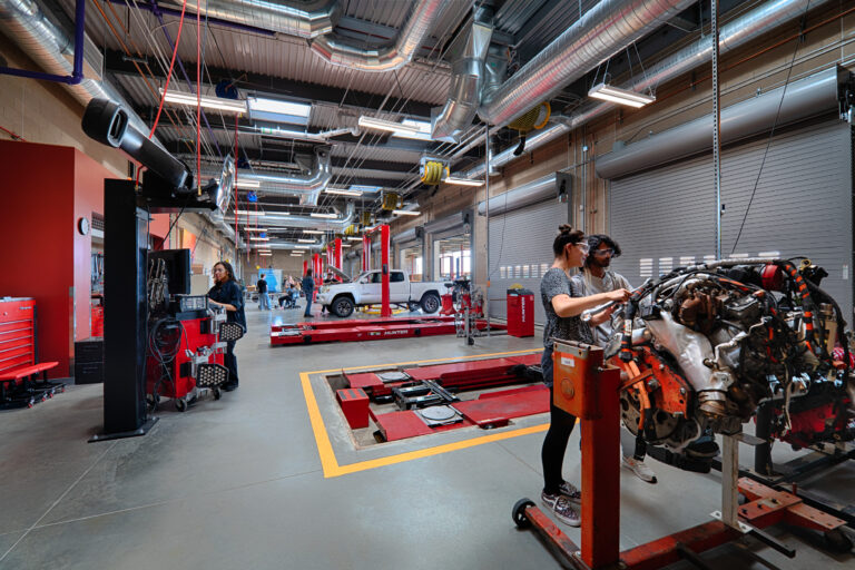 People working in an automotive repair shop with car lifts, a vehicle engine, and a white pickup truck