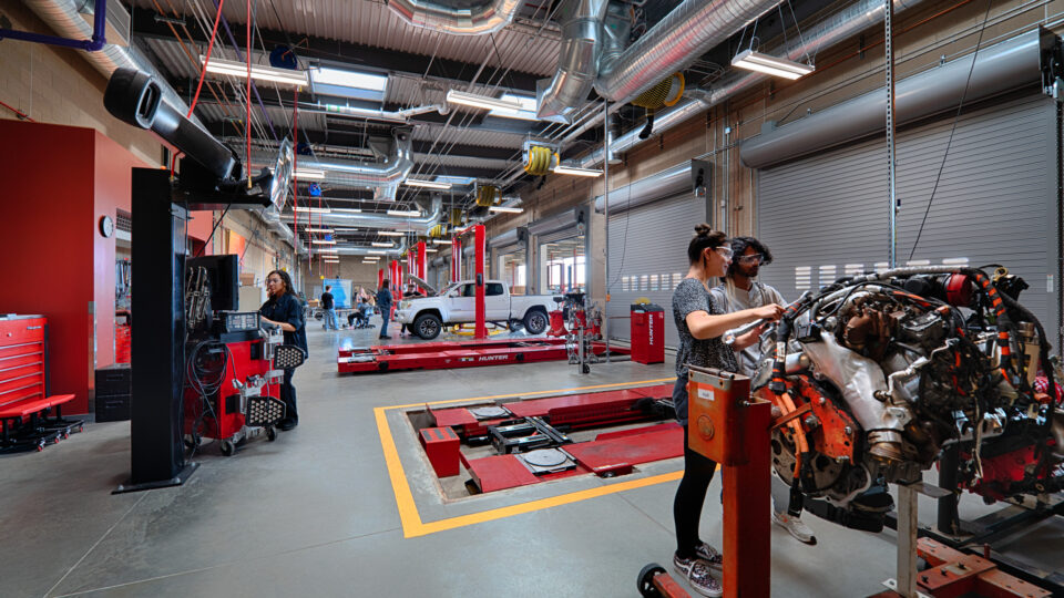 People working in an automotive repair shop with car lifts, a vehicle engine, and a white pickup truck