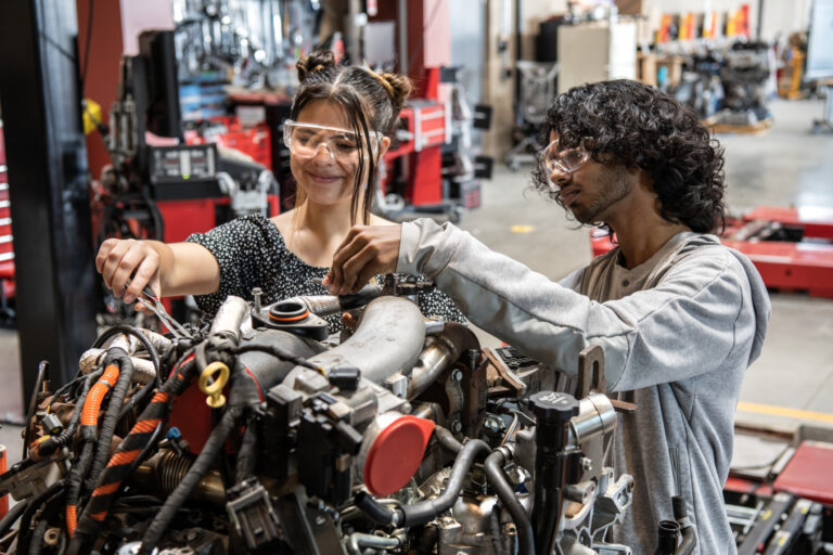 Two students wearing safety goggles work together on a car engine in an auto shop