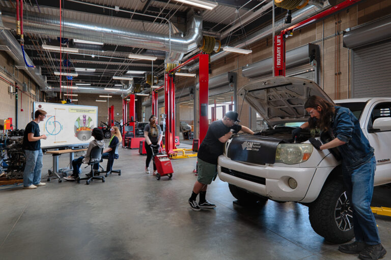 Students and instructors work on cars in a large automotive training garage with lifts and a presentation screen