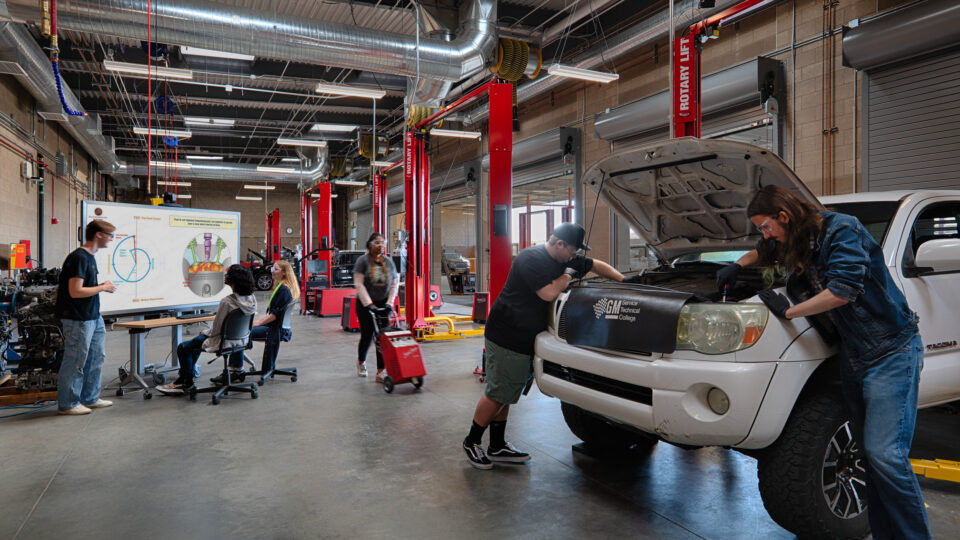 Students and instructors work on cars in a large automotive training garage with lifts and a presentation screen