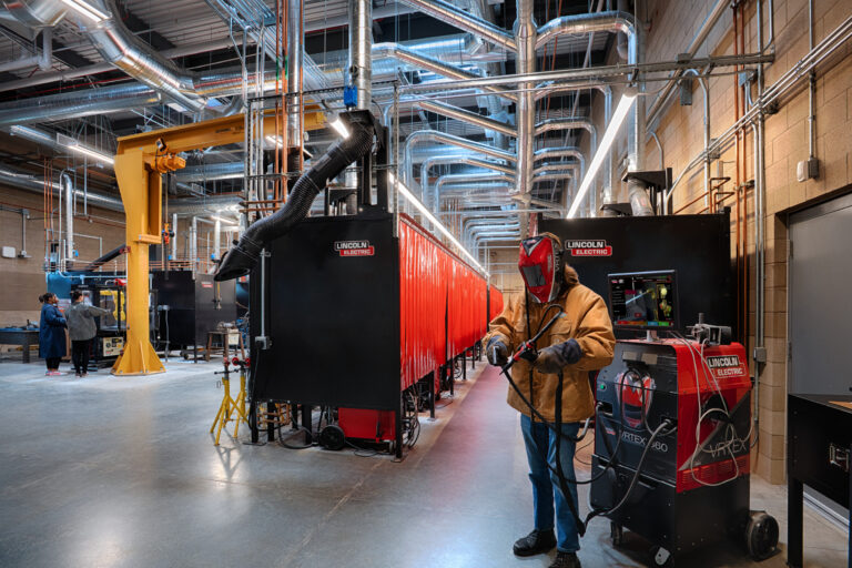 Worker in welding helmet operates a welding torch in a large industrial training lab with Lincoln Electric equipment
