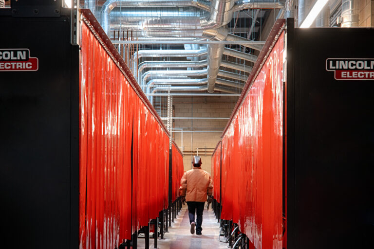 Person walking down a narrow industrial aisle between red welding booths under silver ventilation ducts