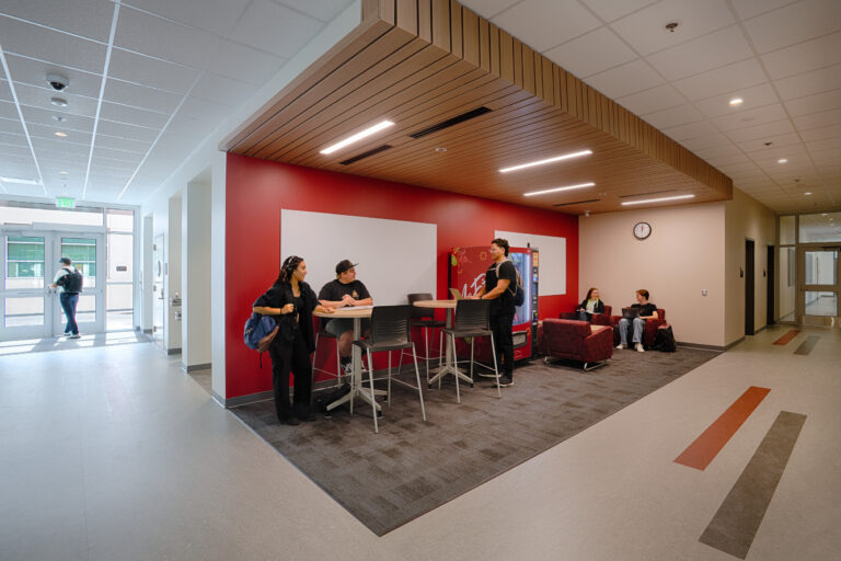 Students socializing in a modern campus hallway lounge with tables, chairs, and red seating area