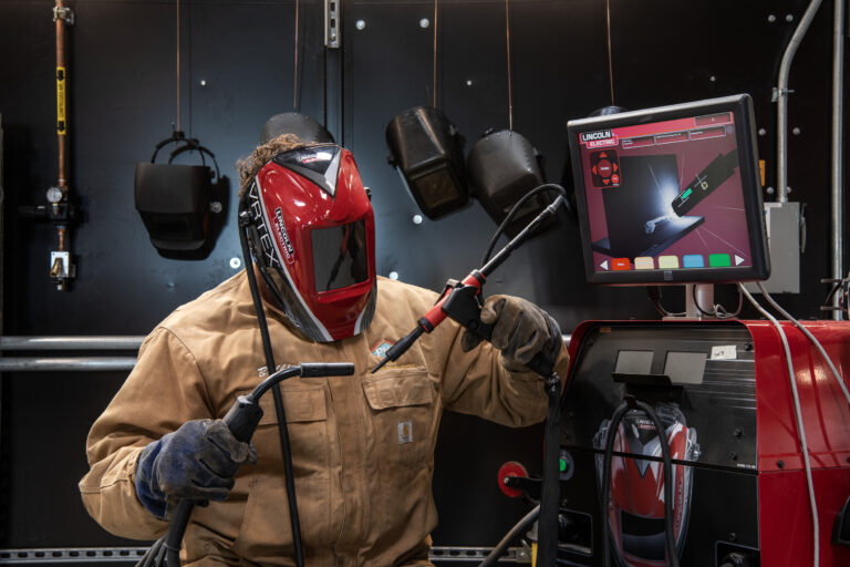 Welded worker in protective gear using a welding torch beside a Lincoln Electric machine and monitor