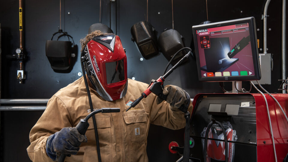 Welded worker in protective gear using a welding torch beside a Lincoln Electric machine and monitor