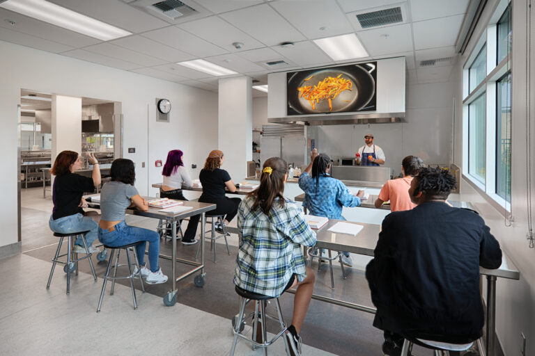Chef demonstrating cooking to students seated at stainless steel tables in a modern culinary classroom