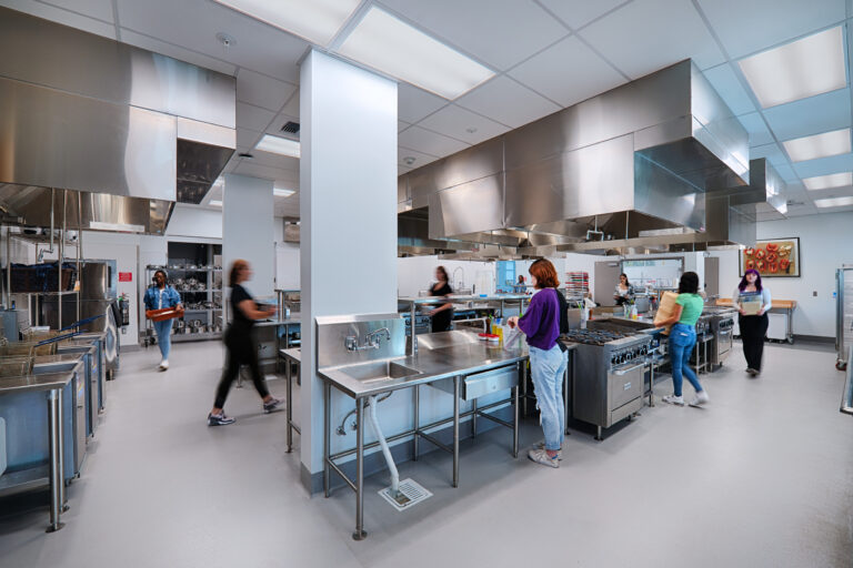 Bright modern commercial kitchen with several people moving between stainless steel workstations and ovens