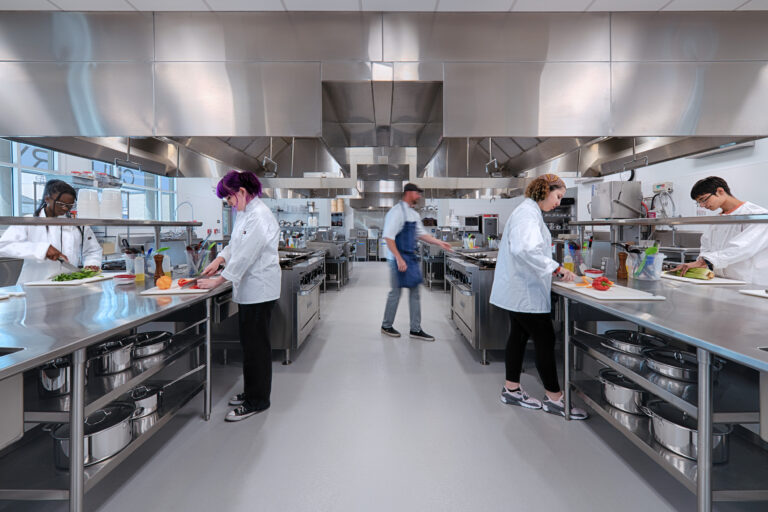Students in white chef coats prepare vegetables at stainless steel counters in a commercial kitchen