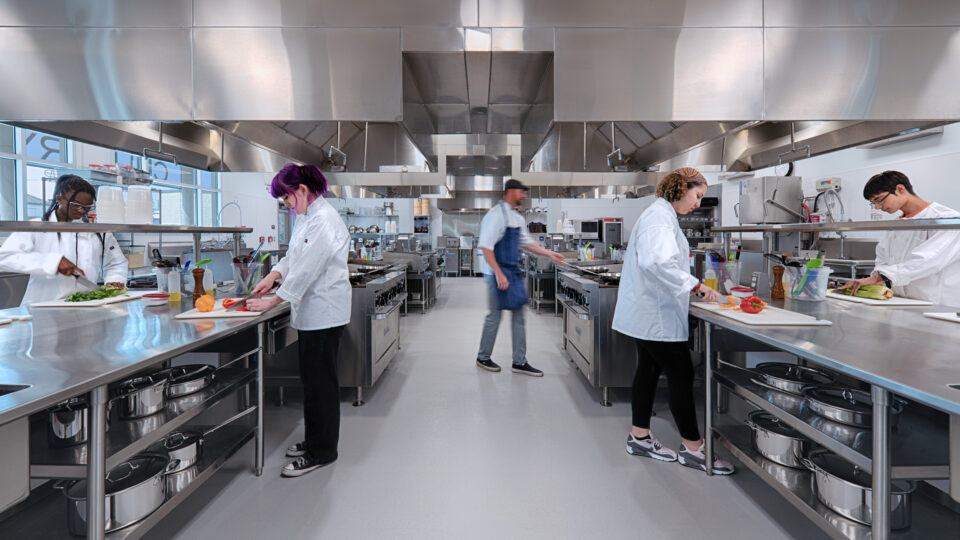 Students in white chef coats prepare vegetables at stainless steel counters in a commercial kitchen