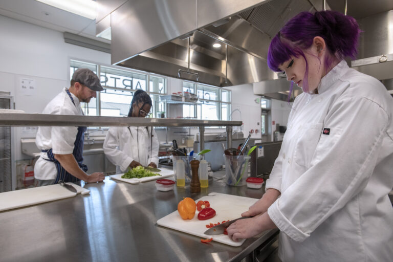 Chefs in white coats prepare vegetables in a professional kitchen, with a purple-haired woman chopping peppers in the foreground