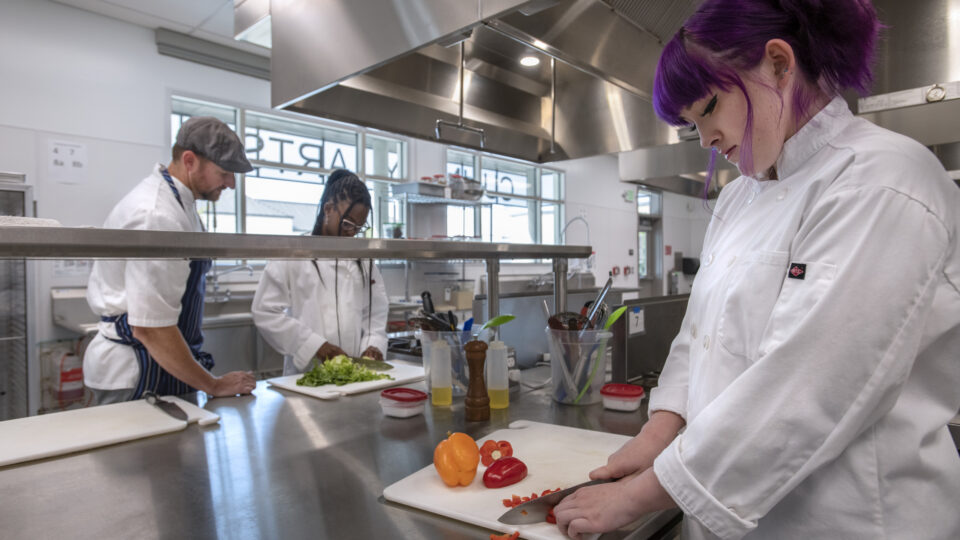 Chefs in white coats prepare vegetables in a professional kitchen, with a purple-haired woman chopping peppers in the foreground