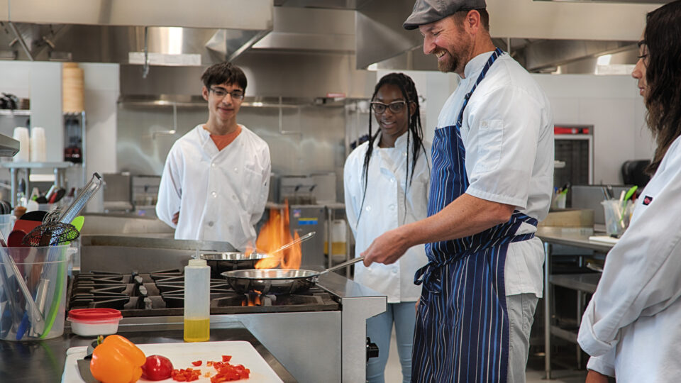 Chef teaching students in a commercial kitchen while cooking at a flaming stove