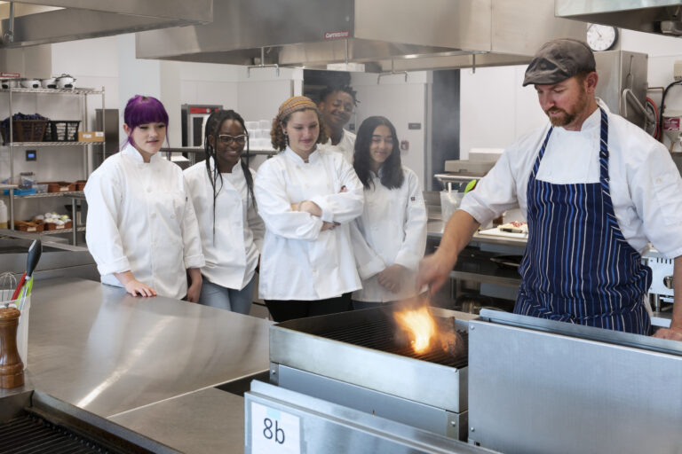 Chef demonstrating a flare-up grill technique to a group of students in a professional kitchen