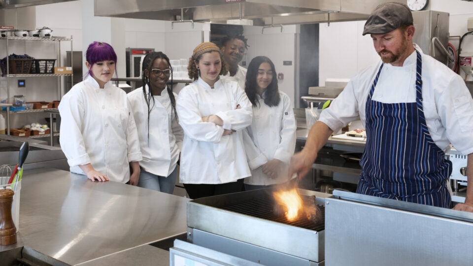 Chef demonstrating a flare-up grill technique to a group of students in a professional kitchen