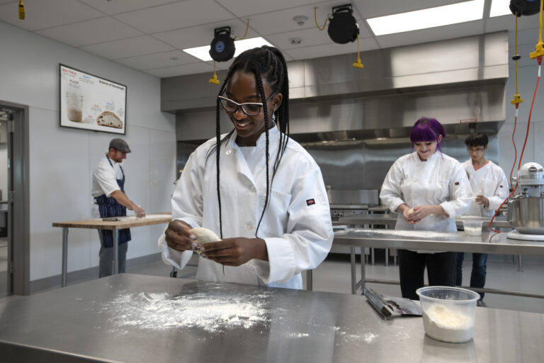 Students in chef coats shaping dough in a professional kitchen classroom