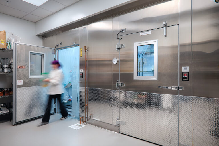 Person in a white coat moving through a stainless steel walk-in cooler with blue strip curtains