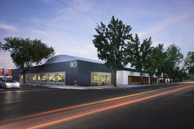 Modern corner building at dusk with large windows, trees, and light trails on a wet street