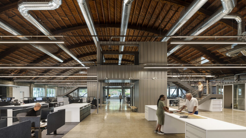 Modern open-plan office with exposed wooden ceiling, large silver ducts, and people talking at a reception desk
