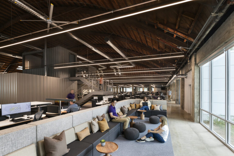 Open-plan industrial office with exposed wood beams, long lights, workstations, lounge seating, and people collaborating