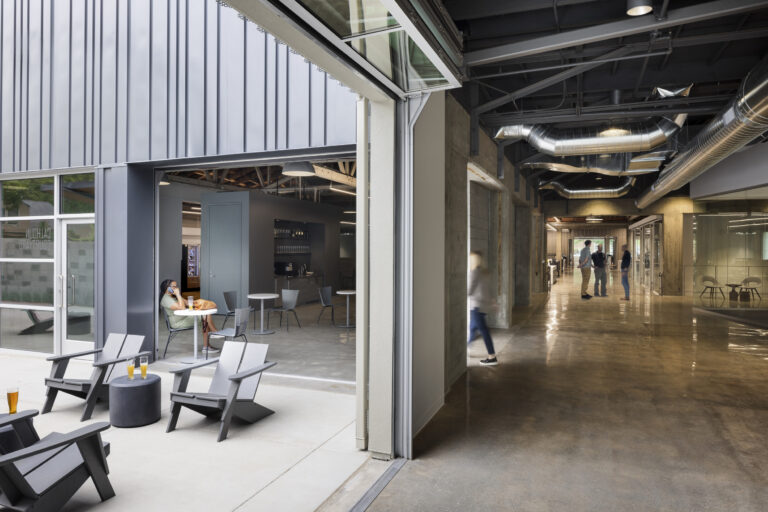 Modern office lounge opening into a hallway with exposed ducts, glass walls, and people chatting inside