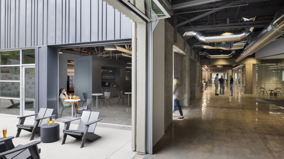 Modern office lounge opening into a hallway with exposed ducts, glass walls, and people chatting inside