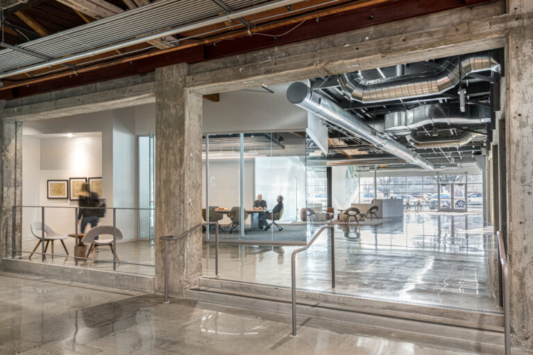 Modern office interior with exposed concrete, glass meeting rooms, and large metal ceiling ducts