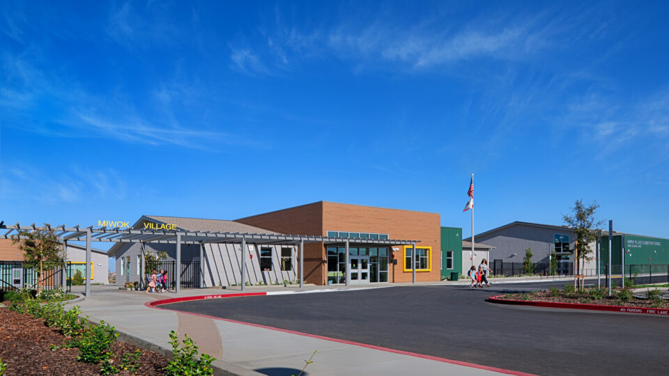 Modern elementary school campus entrance with children walking under a blue sky, labeled Miwok Village Elementary School