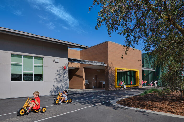 Children riding yellow tricycles outside a modern school building on a sunny day