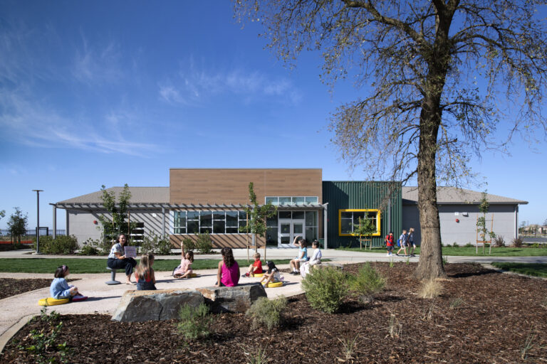 Children sit outside a modern school building while a teacher reads to them on a sunny day