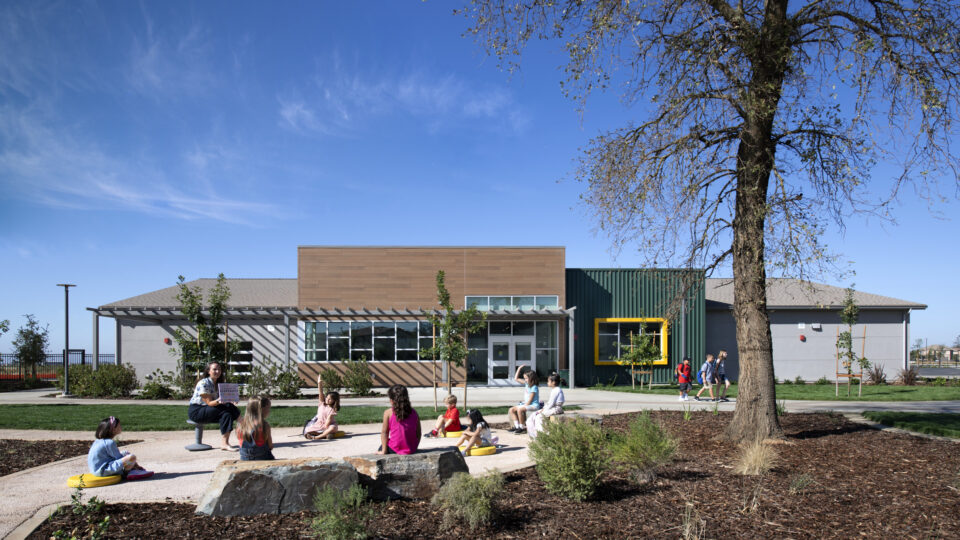 Children sit outside a modern school building while a teacher reads to them on a sunny day