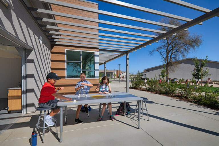 Children playing at an outdoor table under a modern pergola on a sunny school campus