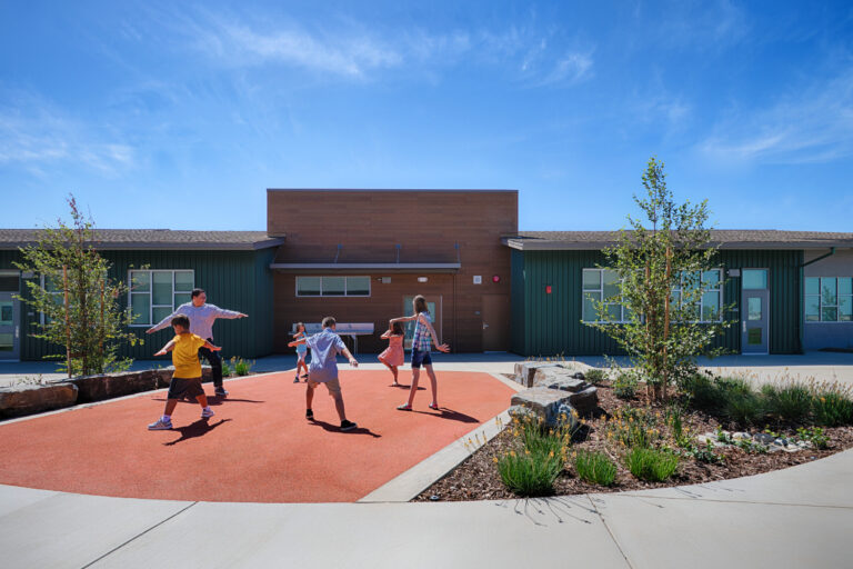 Children and an adult play on a red courtyard at a modern school building under a bright blue sky